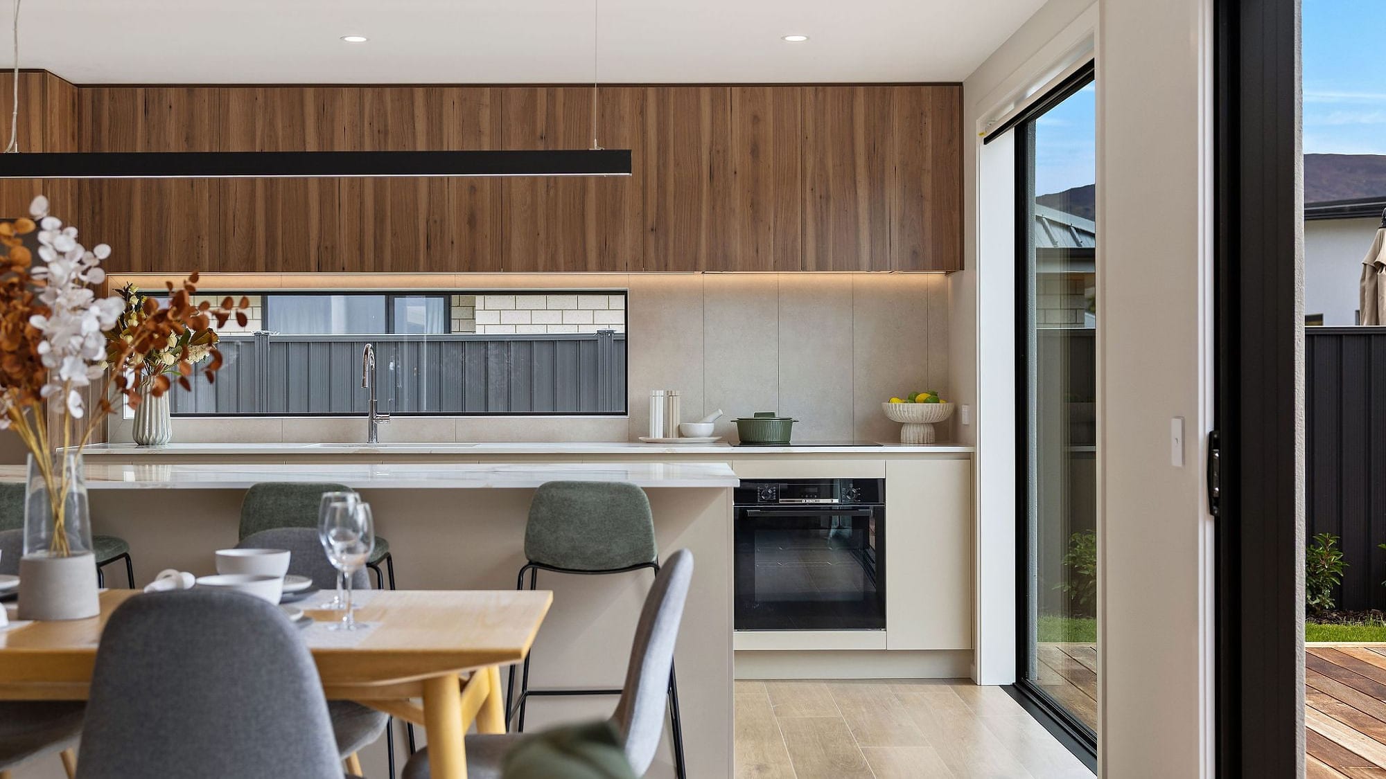 A kitchen splashback in pale large-format stone-look panels, running unbroken between the benchtop and the underside of the timber cabinetry, past a horizontal window and the cooktop.