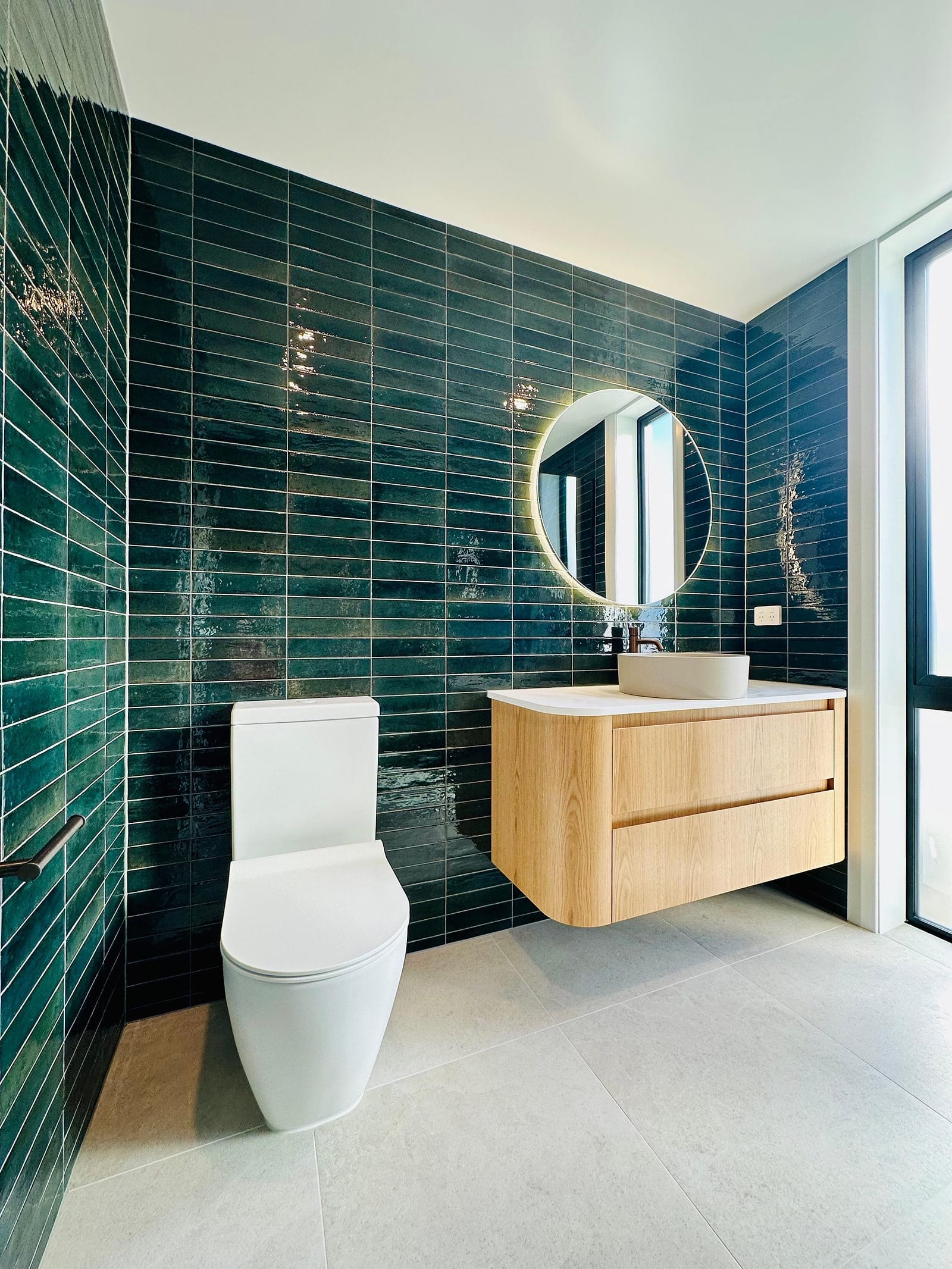 A Bannockburn bathroom in deep green glazed subway tile, stacked in level courses that carry around the internal corner, with a round backlit mirror, an oak vanity and a pale 600×600 porcelain floor.