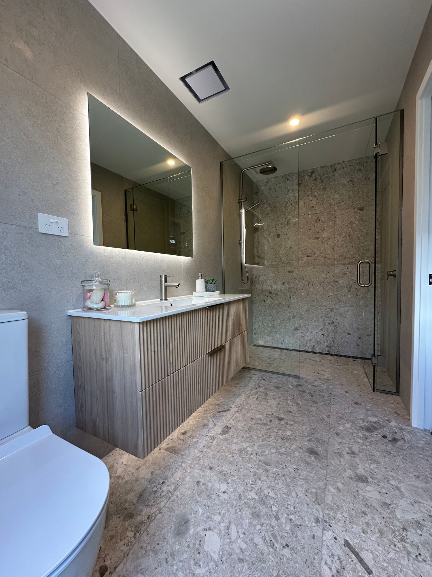A Cromwell bathroom: a fluted vanity under a backlit mirror, a walk-in shower behind glass with a terrazzo-look feature wall, and a channel drain set flush at the shower entry.