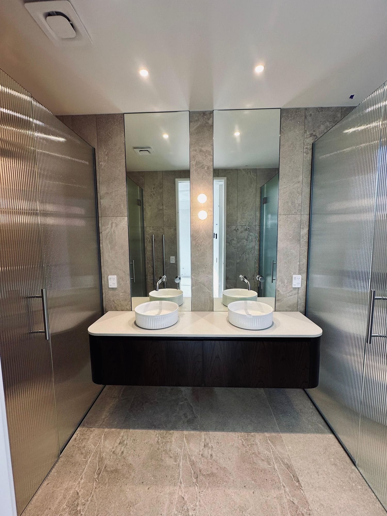 A Wānaka bathroom head on: a fluted tile pilaster centred between two mirrors, stone-look porcelain walls and fluted glass screens either side of the vanity.